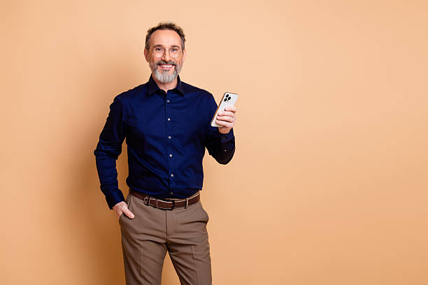 istock Confident mature man holding smartphone against plain beige background while wearing stylish formal attire and smiling 2237093823