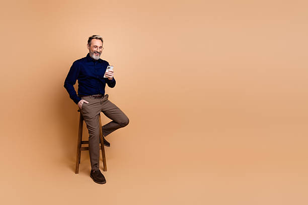 istock Confident mature businessman seated on a stool smiling while holding a smartphone against a plain beige backdrop 2237093894
