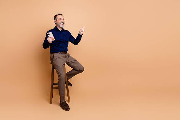 istock Mature businessman holding smartphone while sitting on stool against beige background, pointing confidently with finger 2237093896