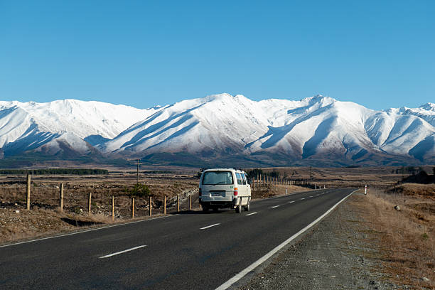 A van heads in the direction of Mount Cook Village, about 50km away, on Mount Cook Road near the junction with State Highway 8 in the southern alps of the South Island.  In the background are mountains in the Mt Cook Range. This image was taken on a cold and sunny morning on 6 September 2025.