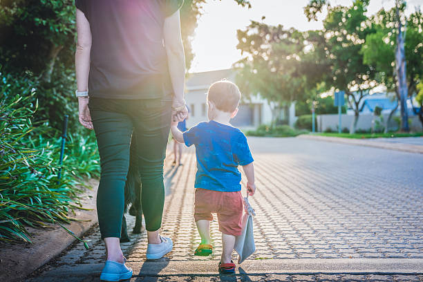 A woman holding hands with a toddler on a paved pathway surrounded by plants in a sunny public park. The setting conveys a warm and peaceful bonding moment captured during a family walk. Soft sunlight filters through the trees, highlighting the togetherness and serenity of the scene.