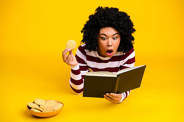 A young woman with curly black hair reacts in surprise while reading a book, holding a cookie, on a vibrant yellow background.