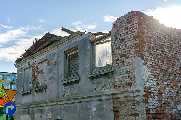 Dilapidated townhouse with crumbling plaster and brick wall, visible damaged windows and partially collapsed roof section.