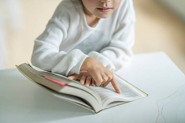 A young child in a white sweater points to text in an open book on a clean white table, illustrating study, learning, and concentration in a calm home or classroom setting.