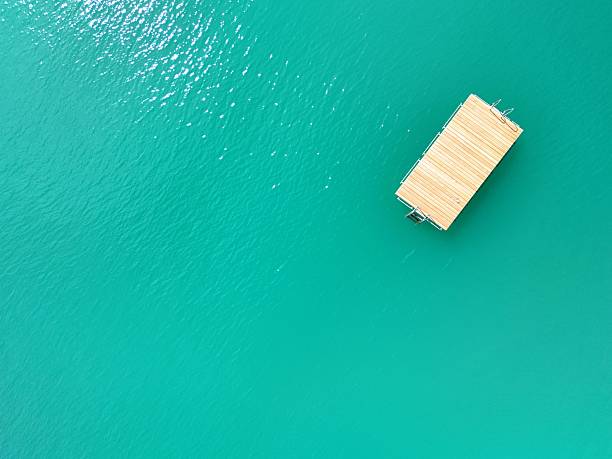 A minimalist aerial photograph of a wooden dock floating on calm, turquoise water. The vibrant color and serene composition create a peaceful, summer-inspired atmosphere. Ideal for travel, nature, and relaxation themes.