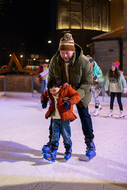 Medium full-length shot of a young boy and his father skating on an ice rink in Newcastle-upon-Tyne, North East England. They are both wearing warm clothing, the boy looking down at his skates while his father supports him.Videos similar to this scenario available.
