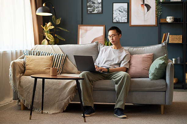 istock Young Adult Asian Man Sitting on Sofa Using Laptop for Remote Work at Home 2237119950
