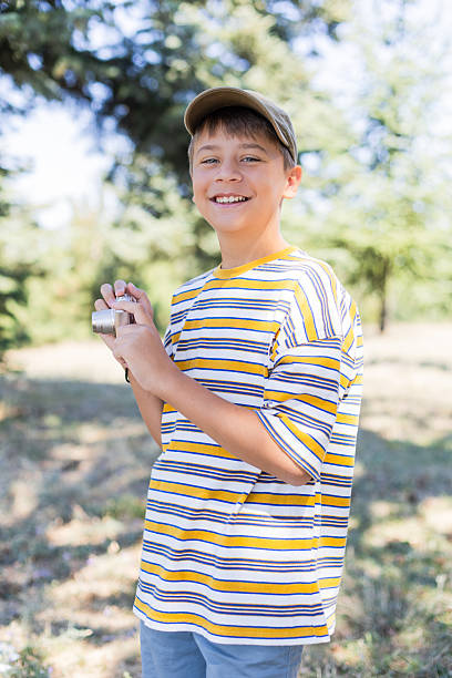 A smiling boy wearing a yellow and white striped T-shirt and a khaki baseball cap stands outdoors. He holds a small silver digital camera in his hands. The background is a bright, sunny, and blurred outdoor setting with trees and greenery. The boy looks directly at the camera with a joyful expression.