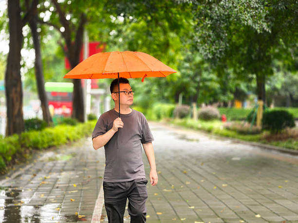 A man was strolling in the rain, accompanied by an orange umbrella