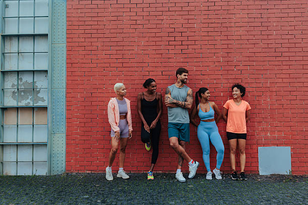 Five multiracial athletes taking a break, leaning against a red brick wall, chatting and smiling after a workout session