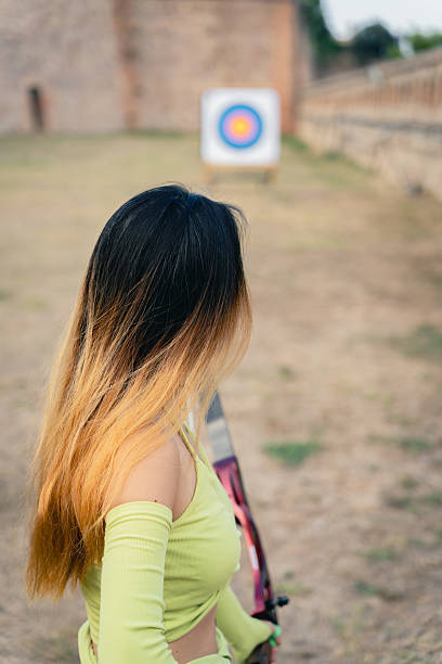 Young woman holding a recurve bow, looking at an archery target in an outdoor setting, focusing on aiming and precision