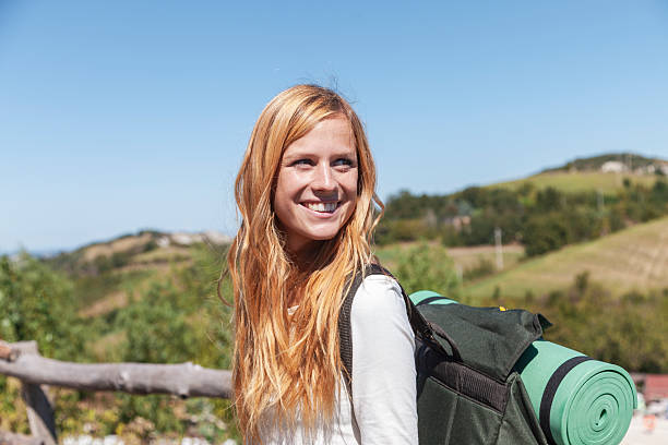 istock Young woman smiling, carrying a backpack and sleeping mat, looking away with hills in background 2237144005