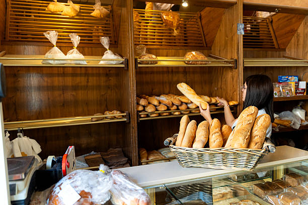 Woman baker working in a traditional micro bakery or bread shop, arranging fresh baguettes on wooden shelves