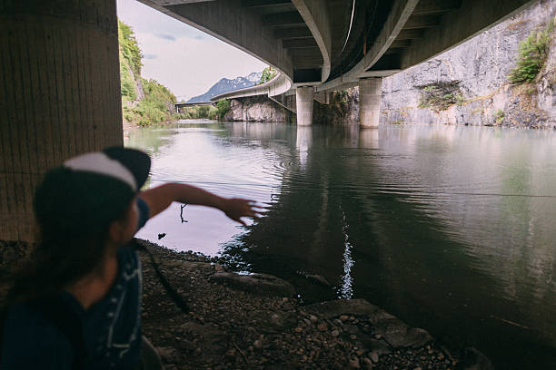 A person joyfully points towards a body of clear water flowing beneath a picturesque bridge, set in a beautiful and natural environment