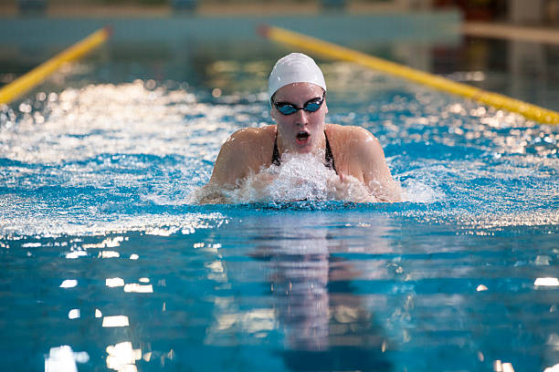 Professional female swimmer in white swim cap and goggles performing breaststroke technique in indoor swimming pool with lane markers. Action shot captures competitive swimming training with water splash and reflection effects. Perfect for competitive sports, swimming training, fitness and athletic performance concepts.