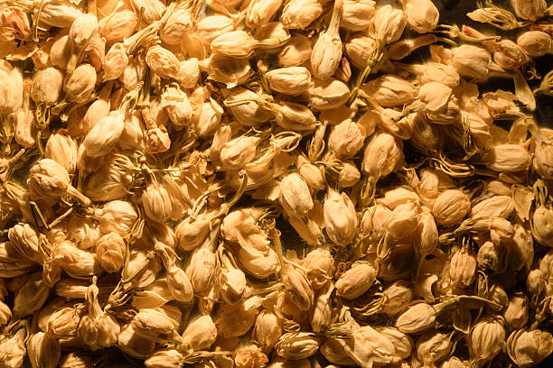 A close-up photograph captures a dense collection of dried jasmine flowers, showcasing their delicate, creamy-yellow petals and stems. The warm lighting enhances their texture and highlights their potential use as an ingredient in aromatic herbal teas and infusions, evoking feelings of calm and natural wellness.