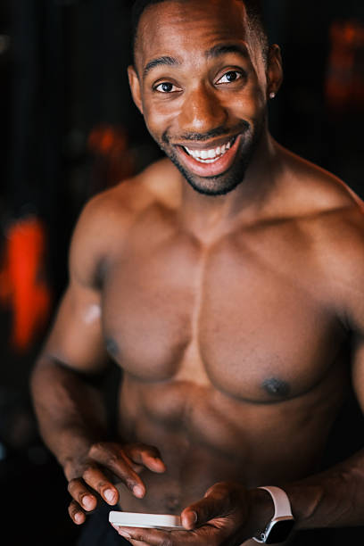 A fit, athletic man sitting shirtless in a modern gym after a workout, smiling and holding a smartphone while resting on a medicine ball. He is wearing black workout shorts, black athletic shoes, and appears relaxed and happy, showing a thumbs-up gesture. Perfect image for fitness, gym lifestyle, workout motivation, health and wellness, athletic training, sports photography, and positive energy themes.
