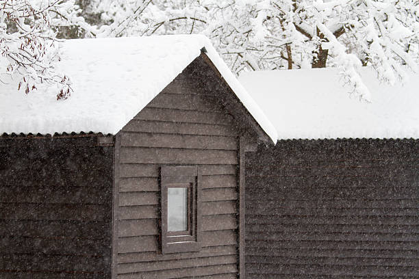 Snowflakes are gently falling down upon a charming wooden house that features a lovely window, highlighting the beauty of winter