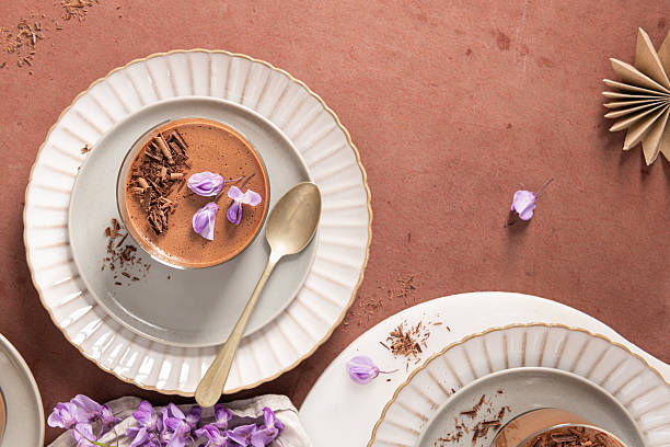 An overhead view featuring two creamy chocolate mousses in glasses, styled on plates with a spoon, flowers, and decorative elements on a rustic surface.