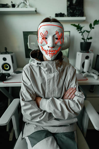 Cyber figure in LED mask stares at camera in minimalistic tech room, embodying hacker lifestyle and cybersecurity awareness with focus on digital protection and system control from home office.