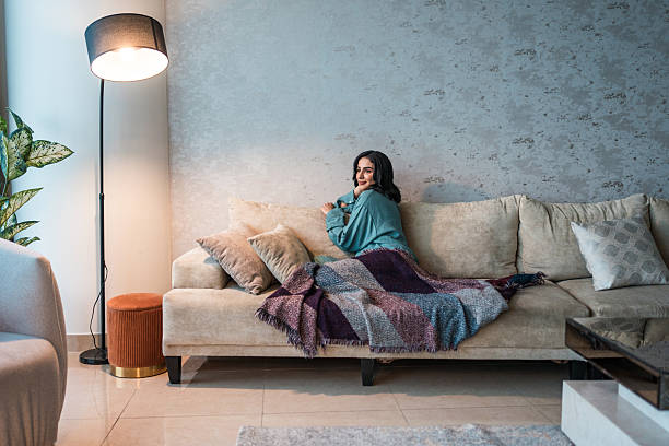 A young Arabic woman enjoys a tranquil moment on a plush sofa wrapped in a colorful blanket in a stylish, modern living room with soft lighting and elegant decor.