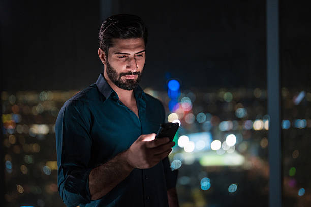 Young Arabic male with a thoughtful expression using a smartphone in a sleek, modern home overlooking the vibrant night cityscape of Riyadh. The setting exudes a sense of technology and connection.