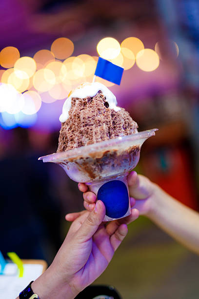 A close-up shot of a large shaved ice dessert being handed from one person to another. The clear plastic bowl holds a mound of shaved ice topped with chocolate crumble and a dollop of cream, all adorned with a small blue flag.