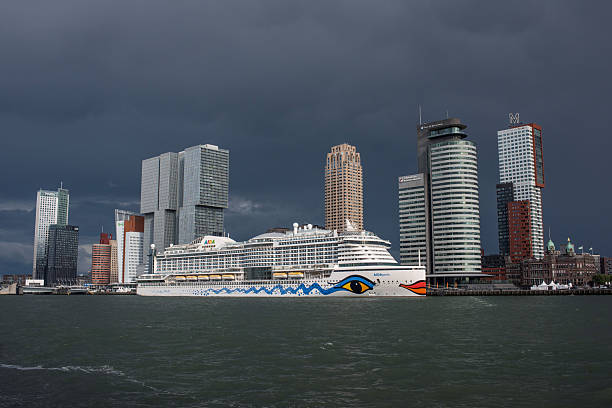The AIDAperla cruise ship is docked along the waterfront in Rotterdam, Netherlands. Modern high-rise buildings of the city’s Kop van Zuid skyline form the backdrop under a dramatic cloudy sky. The scene highlights Rotterdam as an important hub for tourism and the maritime industry