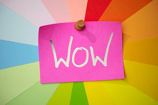 The single word 'WOW' written in a silver marker pink on red note paper, pinned to a colorful array of background blank message notes that radiate around the main message note in a rainbow spiral effect. Conceptual message relating to wow, effect, exclamation, action, excitement, etc.