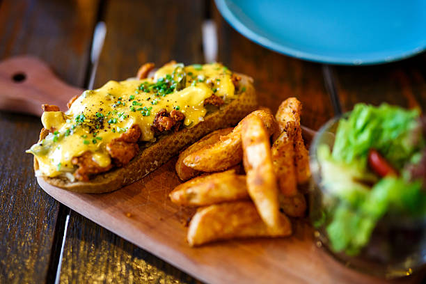 A gourmet open-faced sandwich on a wooden cutting board. The toasted bread is topped with a savory filling and melted yellow cheese, and is served with a generous side of crispy potato wedges and a fresh green salad.