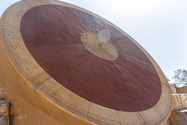 A low-angle close-up shot captures the massive circular face of an ancient astronomical instrument. The disc is divided into a central reddish-brown section and an outer ochre-colored rim with engraved measurement markings and radial lines. The structure, made of stone or stucco, dominates the frame, viewed from a low perspective against a bright, clear sky, emphasizing its geometric design and scientific function in the observatory in Rajasthan.