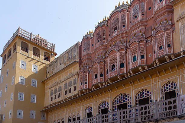 A low-angle, wide shot captures the dramatic contrast in color and style between two sections of a historic palace. On the right, the famous terracotta-pink facade is characterized by rows of ornate jharokha (lattice windows) and domes. On the left, a simpler, ochre-colored tower with small, geometric windows stands adjacent. A carved balustrade runs along the lower level. The vibrant architecture stands out against a bright, pale blue sky in Rajasthan.