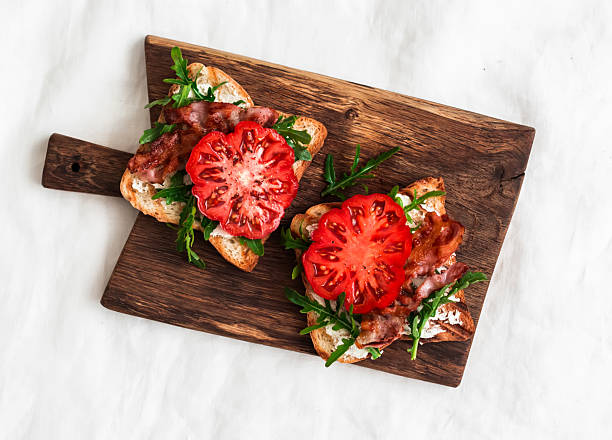 Grilled toasts with cheese, arugula, bacon and tomato - delicious appetizer, tapas, snack, breakfast on a rustic cutting board on a light background, top view