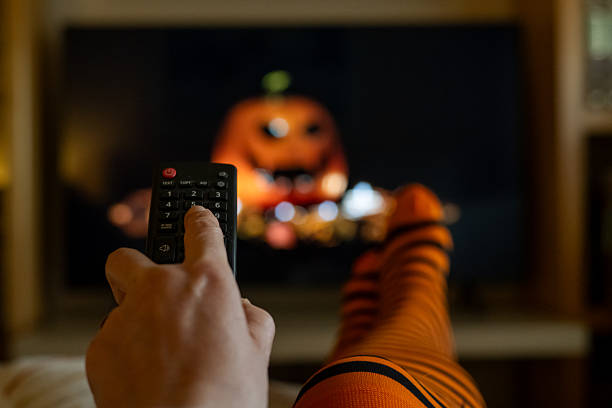 istock Person wearing orange and black striped socks relaxing at home on the sofa, using a remote control for streaming content on a screen displaying a scary pumpkin face during halloween night 2237250175