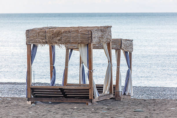 Two beach umbrellas with blue and white curtains. The umbrellas are on the beach next to the ocean