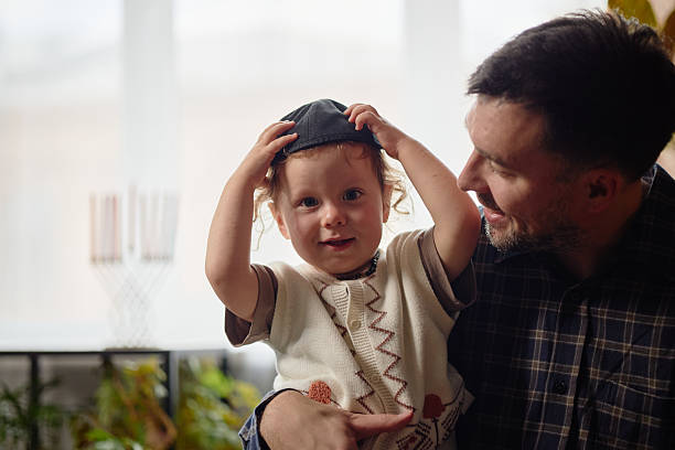 Caucasian man holding toddler adjusting kippah while smiling indoors, menorah visible in background, celebrating Hanukkah together, natural daylight illuminating scene