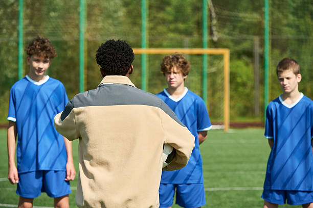 Black man coach instructing group of teenage boys standing on soccer field during outdoor training session, boys listening attentively, goalpost visible in background