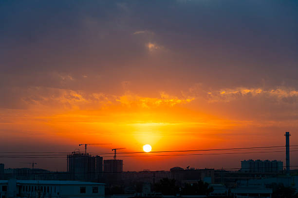 Beautiful city sunset with bright orange and purple sky. Silhouette of buildings and cranes on horizon. Urban evening landscape with dramatic clouds and sunlight