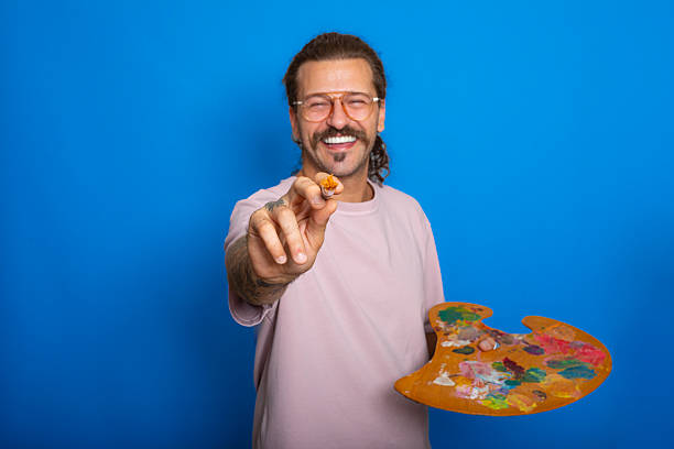 A Caucasian man stands against a wall with a can of paint and a brush.