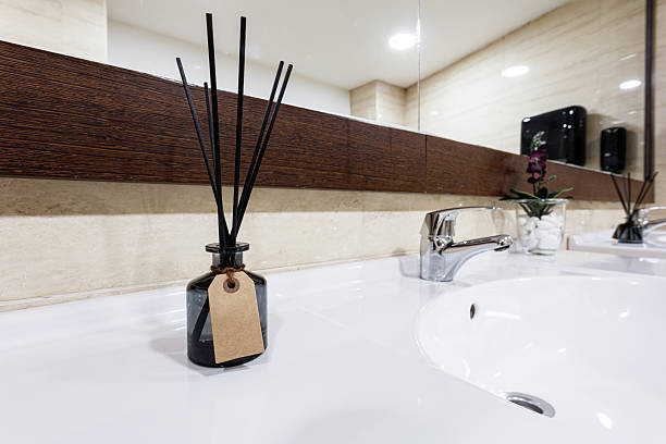A stylish bathroom scene featuring a black diffuser with reed sticks and a blank tag, set on a white counter under a wooden-framed mirror. Subtle decor and plant accents create a calm, spa-like vibe