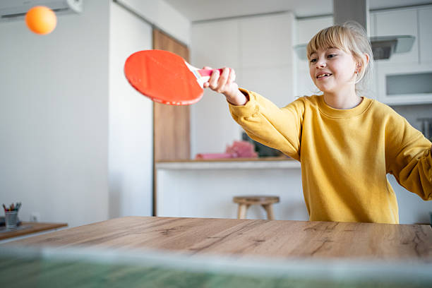 Child in yellow sweatshirt enthusiastically plays table tennis at home, hitting an orange ball with a red paddle while smiling and enjoying active indoor recreation and playtime