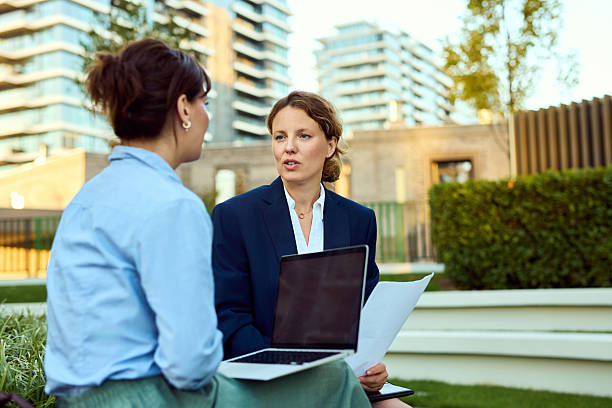 Two Women Business Professionals Discuss Documents Outdoors With Laptop During Meeting