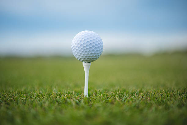 A close-up shot of a golf ball positioned on a tee, surrounded by lush green grass and under a gentle blue sky. The image captures a serene and focused moment in the game of golf, emphasizing tranquility and precision.
