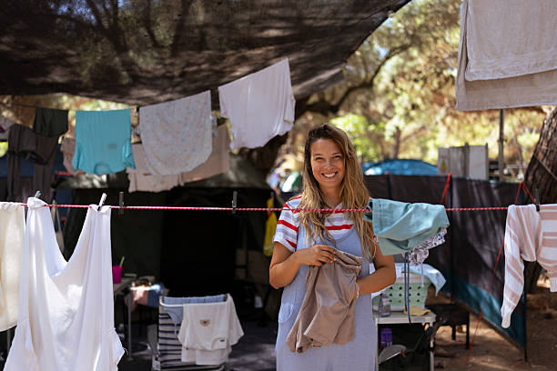 A cheerful woman with long, blonde hair wearing casual attire stands outdoors, hanging laundry on a clothesline in a rustic, sunlit setting. Fabric and trees surround the scene, creating a natural, serene atmosphere.