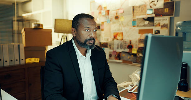 istock Black man, computer and detective in office with research for case, search for lead or problem solving. Mature person, criminal investigation and check surveillance online for crime analysis at night 2237334416