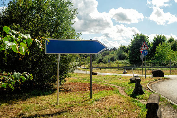 A directional blue road sign standing on the roadside under a bright cloudy sky. Symbol of navigation, guidance, and travel.