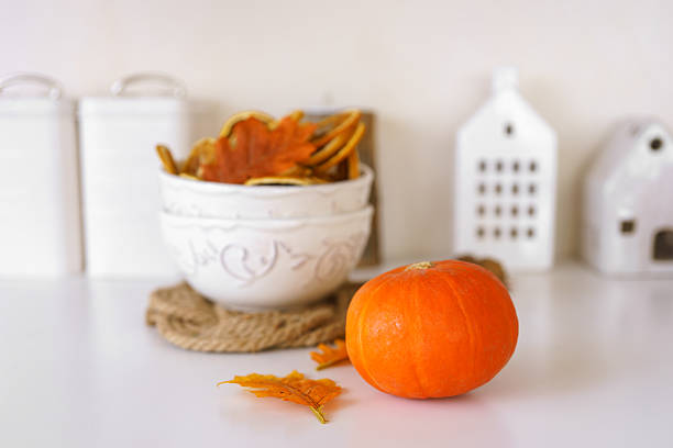 White bowl filled with dried orange slices, orange pumpkin and colorful autumn leaves on background in cozy kithen. Home fall decor, beautiful tableware