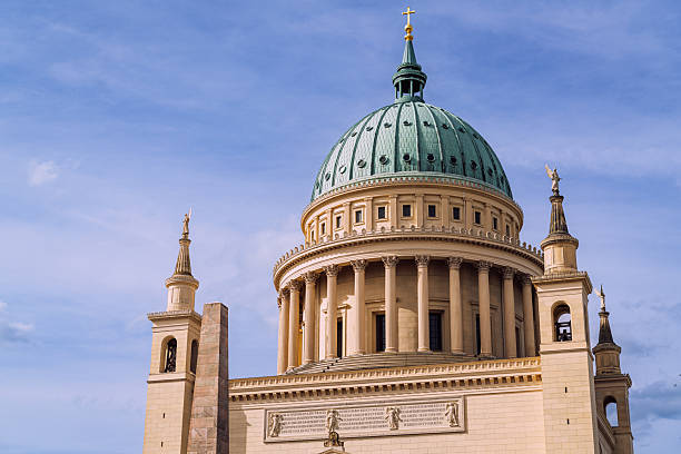 A grand neoclassical building in Potsdam, Germany, crowned with a green dome, flanked by tall columns and statues. Captured against a clear blue sky to convey majesty, history, and cultural heritage.