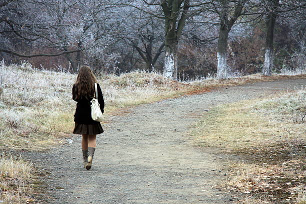 Young woman with long hair and a white bag walks alone on a dirt path through frosty trees in late autumn, creating a quiet, contemplative mood in a cold natural landscape
