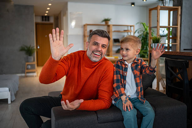 Smiling father and young son sitting on a sofa, waving cheerfully at the camera during a casual video communication, connecting with family or friends while staying at home